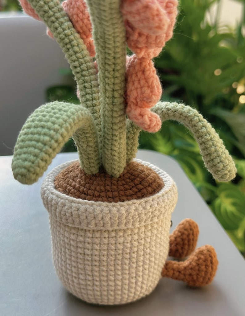Puede incluir: Planta de ganchillo hecha a mano en una maceta blanca con tierra marr&oacute;n. La planta tiene tallos y hojas verdes y flores rosas. La maceta tiene peque&ntilde;os pies marrones. El dise&ntilde;o es caprichoso.