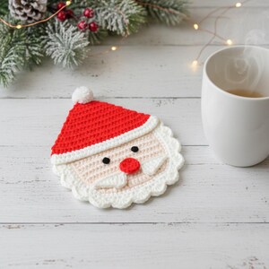 May include: A handmade crochet Santa Claus coaster with a red hat, white beard, and black eyes. The coaster is placed on a white wooden surface, next to a white mug. Festive holiday decor.