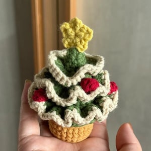 Könnte beinhalten: Ein handgehäkelter Weihnachtsbaum mit gelbem Stern und roten Akzenten. Der Baum ist grün mit cremefarbenen Verzierungen und steht in einem kleinen, gelb-orangenen Topf. Eine festliche Dekoration.