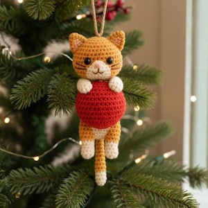 May include: A handmade crochet cat ornament in shades of orange, white, and red, hanging from a Christmas tree. The cat is holding a red ball. The ornament has black eyes and a small pink nose. The cat's tail is hanging down.