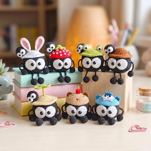 May include: A collection of seven small, round, black crocheted creatures with large white eyes. Each figure wears a unique crocheted hat, including designs like a white bunny ear hat, a red mushroom cap, a green frog hat, and a brown sun hat.