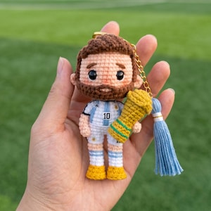 May include: A crocheted amigurumi keyring featuring a figure with brown hair and a beard, wearing a white and light blue football jersey with the number 10. The figure holds a small gold-coloured trophy and includes a light blue yarn tassel.