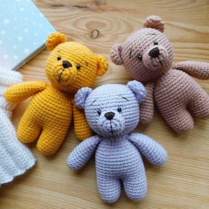 Op de afbeelding: Drie gehaakte teddyberen in verschillende kleuren: geel, paars en bruin. De beren zitten op een houten oppervlak.
