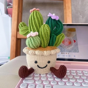 May include: A crocheted cactus plant in a beige pot with a smiling face and brown heart-shaped feet. The cactus has green and dark green sections with pink and purple flower accents. The pot is sitting on a pink keyboard.