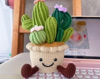 May include: A crocheted cactus plant in a beige pot with a smiling face and brown heart-shaped feet. The cactus has green and dark green sections with pink and purple flower accents. The pot is sitting on a pink keyboard.