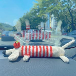 May include: A handmade crochet dachshund dog toy. The toy features a red and white striped body, cream-colored legs and head, and a small red hat. The toy is lying on a gray surface.