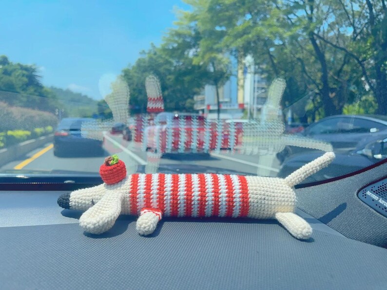 May include: A handmade crochet dachshund dog toy. The toy features a red and white striped body, cream-colored legs and head, and a small red hat. The toy is lying on a gray surface.