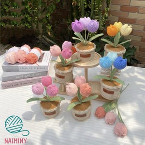 Op de afbeelding: Een verzameling handgemaakte gehaakte tulpen in verschillende kleuren, waaronder roze, paars, oranje, geel en blauw. De tulpen staan in kleine, geweven potten met een decoratieve rand. Sommige staan op een houten standaard, en bollen wol zijn zichtbaar op de achtergrond.