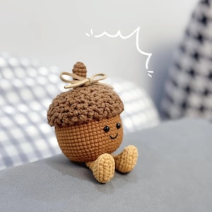 Könnte beinhalten: Eine braune gehäkelte Eichel mit einem lächelnden Gesicht und zwei kleinen Beinen. Die Eichel sitzt auf einem grauen Sofa.