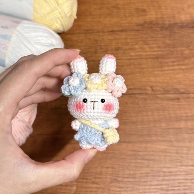 May include: A white crocheted bunny with pink cheeks wearing a flower crown with blue, yellow, and pink flowers. The bunny is wearing a light blue dress and a yellow crocheted bag.