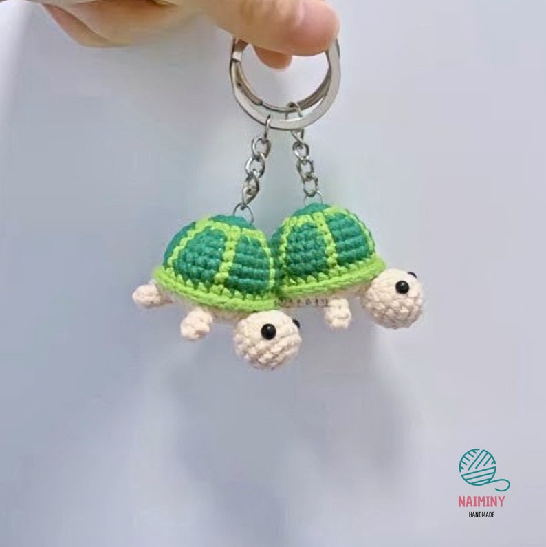 Op de afbeelding: Twee gebreide schildpad sleutelhangers met groene schalen en witte lichamen. De sleutelhangers zijn bevestigd aan een zilveren sleutelring.
