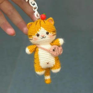 May include: A crocheted orange and white tabby cat keyring featuring a small pink fish-shaped crossbody bag, a tiny red bead accent at the top, and a white metal clasp. The cat has embroidered black eyes, a pink nose, and blush-coloured cheeks.