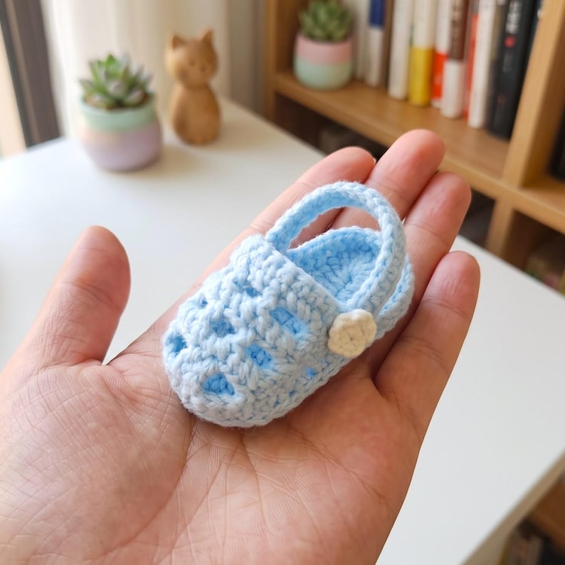 Op de afbeelding: Een kleine, lichtblauwe gehaakte babysandaal met een uitgesneden ontwerp op de bovenkant, een ronde neus en een beweegbare hielband die aan de zijkant is vastgezet met een klein, wit bloemetje met vijf bloemblaadjes.
