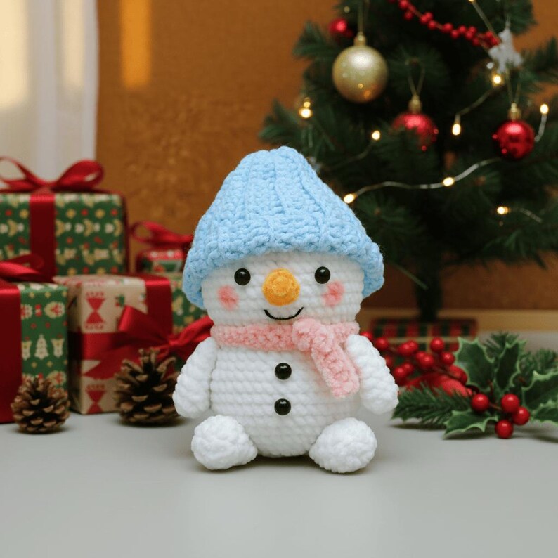 May include: A crocheted snowman toy, wearing a light blue hat and a pink scarf. The snowman has a carrot nose, rosy cheeks, and black button eyes. It is a festive decoration, ideal for the holiday season. Presents and a Christmas tree are in the background.