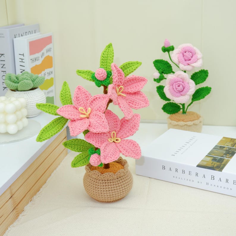 May include: Crocheted pink flowers with green leaves in a brown pot. A second crocheted flower arrangement with pink roses is also visible. The flowers are on a white surface with books in the background.