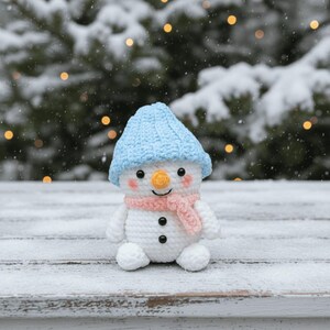 May include: A crocheted snowman figure wearing a light blue hat and a pink scarf. The snowman has a carrot nose, black button eyes, and a smiling mouth. The snowman is sitting on a snow-covered surface.