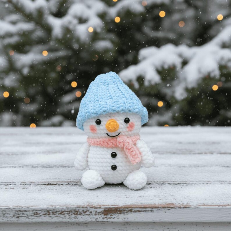 May include: A crocheted snowman figure wearing a light blue hat and a pink scarf. The snowman has a carrot nose, black button eyes, and a smiling mouth. The snowman is sitting on a snow-covered surface.