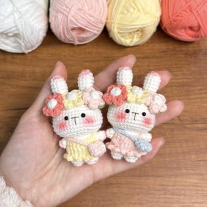May include: Two crocheted bunny plushies with floral headbands. The bunny on the left is wearing a yellow dress and the bunny on the right is wearing a pink dress.