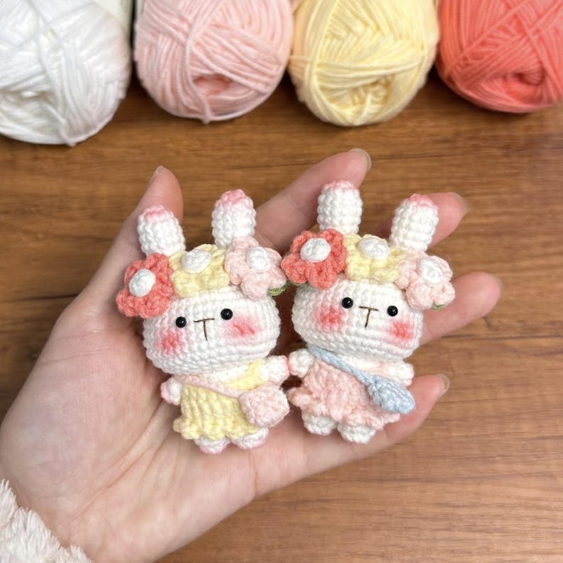 May include: Two crocheted bunny plushies with floral headbands. The bunny on the left is wearing a yellow dress and the bunny on the right is wearing a pink dress.