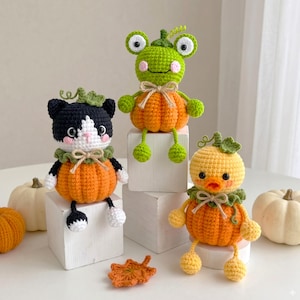 May include: Three handmade crochet plushies featuring a black and white cat, a green frog, and a yellow duck, each sitting on an orange pumpkin base with a green stem, a ruffled collar, and a small beige bow, displayed on white wooden blocks.