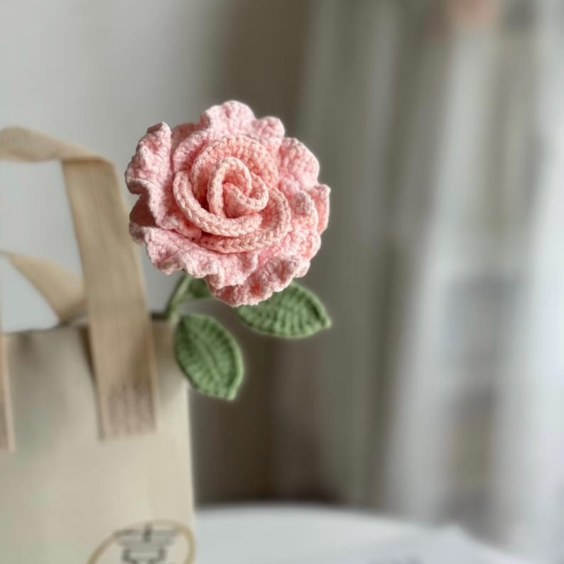 May include: A crocheted pink rose with green leaves. The rose is a handmade craft item.