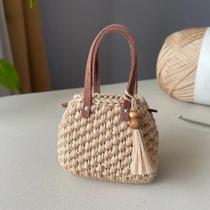 May include: A small, beige crocheted handbag with brown leather handles and accents. The bag features a tassel and wooden bead detail. The bag is approximately 10 cm wide and 7.5 cm tall.