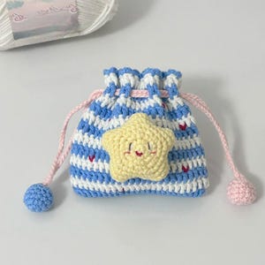 Op de afbeelding: Een handgemaakt gehaakt trekkoordzakje in blauw-witte strepen, versierd met een gele sterapplicatie. Het zakje heeft roze trekkoorden en blauwe en roze pompons. Kleine rode hartjes zijn verspreid over de blauw-witte strepen.