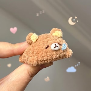 Puede incluir: Una pequeña cabeza de oso tejida a ganchillo en color marrón claro con textura difusa. El oso tiene orejas interiores de color crema, ojos cerrados bordados en hilo marrón oscuro, hocico blanco con nariz marrón oscuro y una gota azul claro.