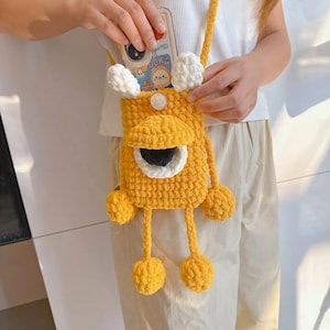 May include: A mustard yellow crochet crossbody phone pouch featuring a large central eye with a black pupil and white iris, two white tufted ears at the top, and two dangling arms with round hands. The pouch has a long matching crochet strap.