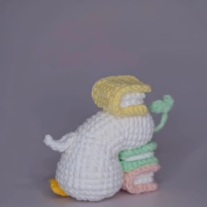 K&ouml;nnte beinhalten: Handgefertigte H&auml;kelentenfigur in Wei&szlig;, Gelb, Gr&uuml;n und Rosa. Die Ente tr&auml;gt eine gelbe Haube und h&auml;lt einen Stapel bunter B&uuml;cher. Die Ente hat einen gelben Schnabel und gelbe F&uuml;&szlig;e. Die Figur steht vor grauem Hintergrund.