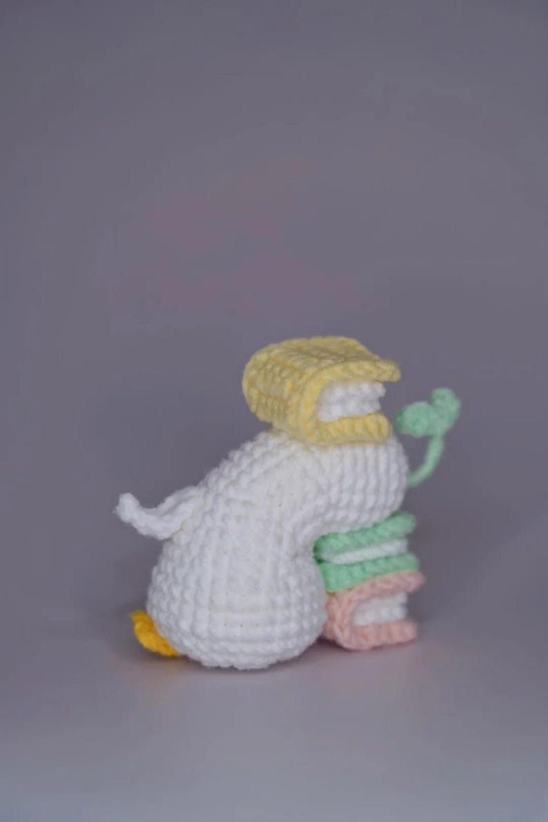 K&ouml;nnte beinhalten: Handgefertigte H&auml;kelentenfigur in Wei&szlig;, Gelb, Gr&uuml;n und Rosa. Die Ente tr&auml;gt eine gelbe Haube und h&auml;lt einen Stapel bunter B&uuml;cher. Die Ente hat einen gelben Schnabel und gelbe F&uuml;&szlig;e. Die Figur steht vor grauem Hintergrund.