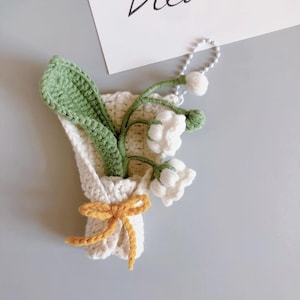 Könnte beinhalten: Ein gehäkelter Strauß weißer Maiglöckchen mit grünen Blättern, gebunden mit einem gelben Band. Der Strauß befindet sich in einem weißen gehäkelten Beutel und hat eine silberne Kette.