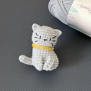 May include: A small crocheted grey cat with a yellow collar. The cat is sitting with its tail curled around its legs.