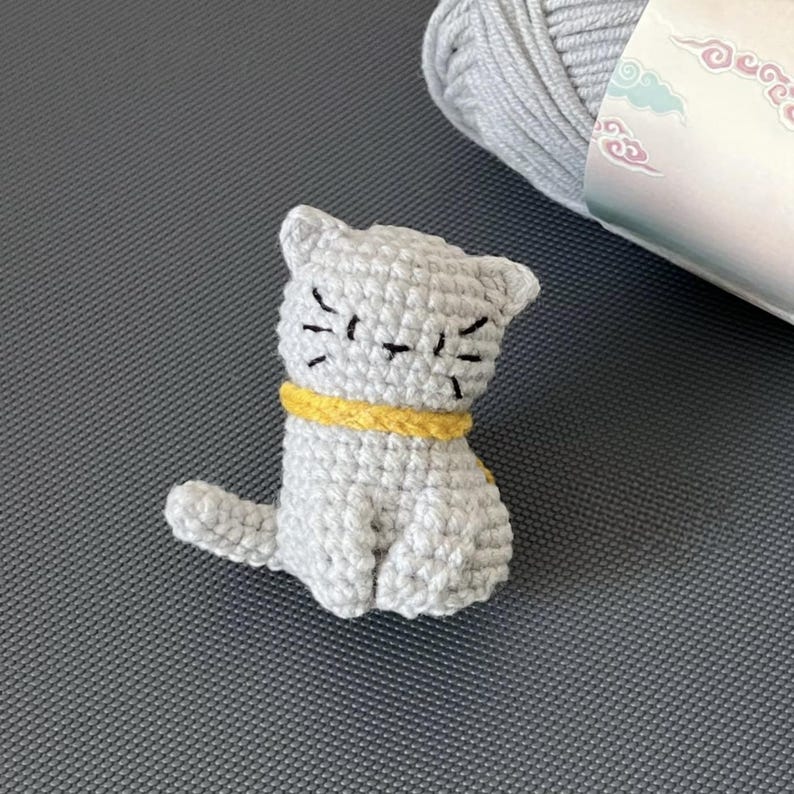 May include: A small crocheted grey cat with a yellow collar. The cat is sitting with its tail curled around its legs.