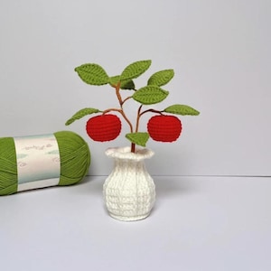 Op de afbeelding: Een handgemaakte gehaakte appelboom met rode appels en groene bladeren in een witte gehaakte vaas. Een bol groen garen staat op de achtergrond. De boom is een decoratief item.