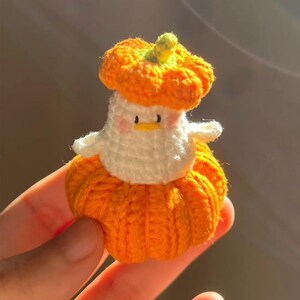 May include: A handmade crochet duck wearing a pumpkin costume. The duck is white with a yellow beak and pink cheeks. The pumpkin costume is orange with a green stem. The duck is small and appears to be a decorative item.