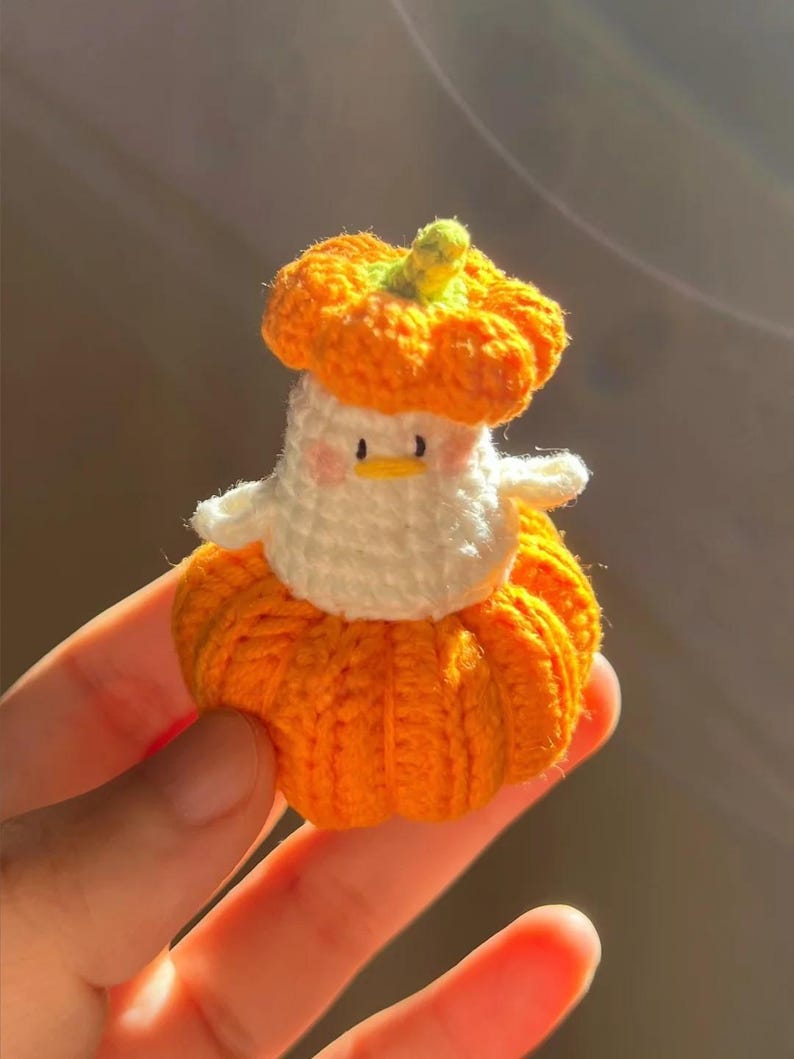 May include: A handmade crochet duck wearing a pumpkin costume. The duck is white with a yellow beak and pink cheeks. The pumpkin costume is orange with a green stem. The duck is small and appears to be a decorative item.