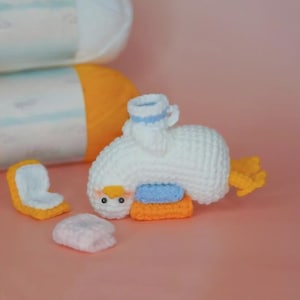 May include: A white crocheted duck toy with a yellow beak and orange feet, featuring a small blue and white hat. The duck is positioned next to a small orange and white crocheted square and a light pink crocheted piece, with yarn rolls behind.
