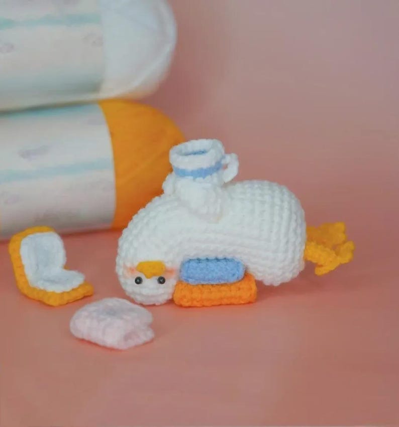 May include: A white crocheted duck toy with a yellow beak and orange feet, featuring a small blue and white hat. The duck is positioned next to a small orange and white crocheted square and a light pink crocheted piece, with yarn rolls behind.