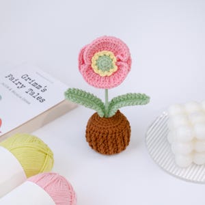Puede incluir: Una flor de ganchillo hecha a mano en una maceta marrón. La flor tiene pétalos rosas con un centro amarillo y verde. Las hojas verdes se extienden desde el tallo. Un libro titulado "Grimm's Fairy Tales" está en el fondo.