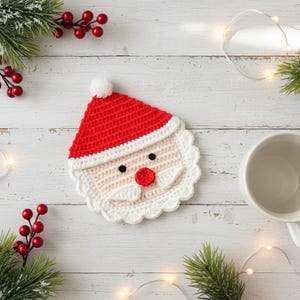 May include: A handmade crochet Santa Claus coaster. The coaster features a red hat with a white pom-pom, a white beard, and a pink face with black eyes and a red nose. The coaster is surrounded by white trim.