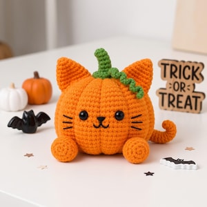 Puede incluir: Una calabaza naranja tejida a ganchillo con forma de gato, con un tallo verde, ojos de cuentas negras y bigotes bordados. La figura tiene dos orejas puntiagudas, dos patas delanteras redondas y una cola rizada. Un letrero de madera dice TRICK OR TREAT.