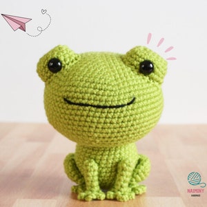 May include: A green crocheted frog amigurumi toy with black eyes. The frog is sitting on a wooden surface.