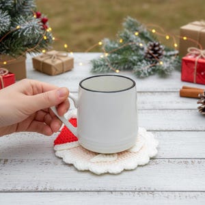 May include: A white ceramic mug with a black rim rests on a Santa Claus-themed crochet coaster. The coaster features red and white yarn, with a scalloped edge. The background includes Christmas decorations and wrapped gifts.