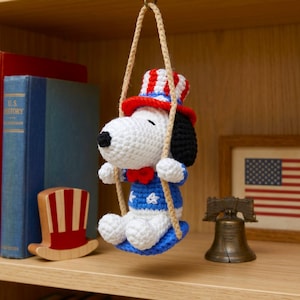 Könnte beinhalten: Eine gehäkelte Snoopy-Figur mit einem rot-weiß-blauen Zylinder und einem passenden blauen Hemd mit weißen Sternen und einer roten Fliege. Die Figur sitzt auf einer blauen, gehäkelten Schaukel, die an einem beigen Seil hängt und einen weißen Körper hat.