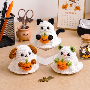 Op de afbeelding: Drie gehaakte spookfiguren van wit garen, elk met een kleine oranje pompoen. Eén spook heeft zwarte kattenoren en een staart, één heeft bruine hondenoren en één heeft groene kikkerkenmerken. Elk figuurtje heeft zwarte kraaloogjes en een mond.