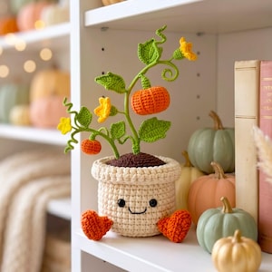 May include: A crocheted pumpkin plant in a cream-colored pot with a smiling face, black eyes, and orange feet. The plant features green stems, leaves, and curly vines with small yellow flowers and two orange pumpkins attached to the stems.