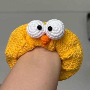 May include: A yellow crocheted headband with large white eyes and a small orange beak, resembling a cartoon bird. The headband is made of textured yarn and is worn on the head.