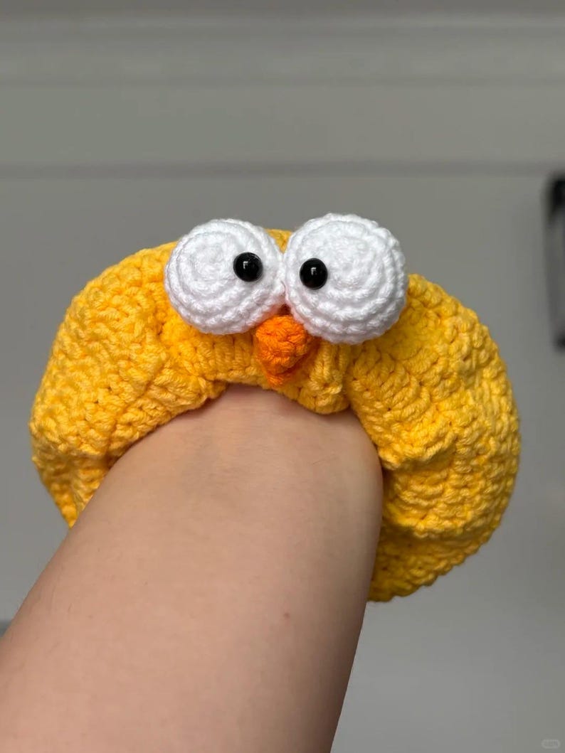 May include: A yellow crocheted headband with large white eyes and a small orange beak, resembling a cartoon bird. The headband is made of textured yarn and is worn on the head.
