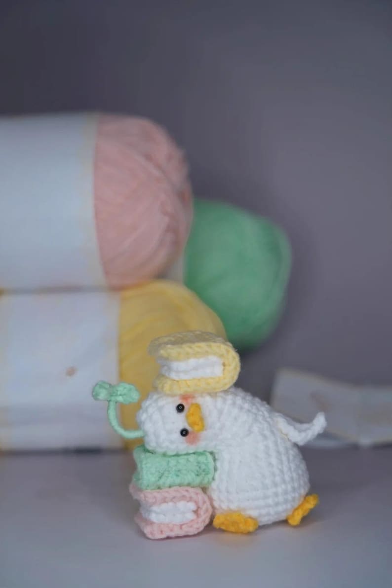 K&ouml;nnte beinhalten: Eine handgefertigte wei&szlig;e H&auml;kelente mit gelbem Hut, die bunte Macaron-f&ouml;rmige Gegenst&auml;nde tr&auml;gt. Die Ente hat orangefarbene F&uuml;&szlig;e und einen gr&uuml;nen Stiel. Im Hintergrund befinden sich pastellfarbene Wollkn&auml;uel.